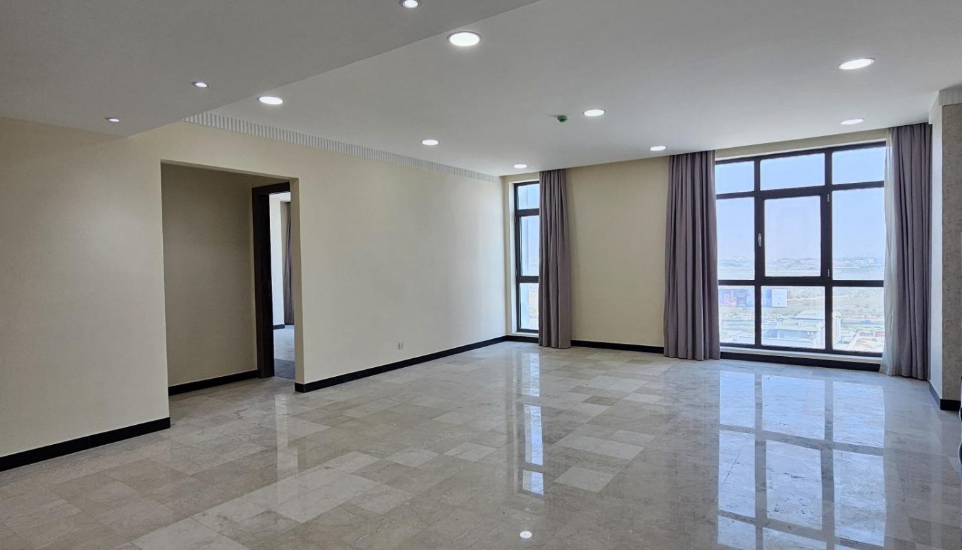 A spacious, empty room with tiled floors, large windows with curtains, beige walls, and recessed ceiling lights.