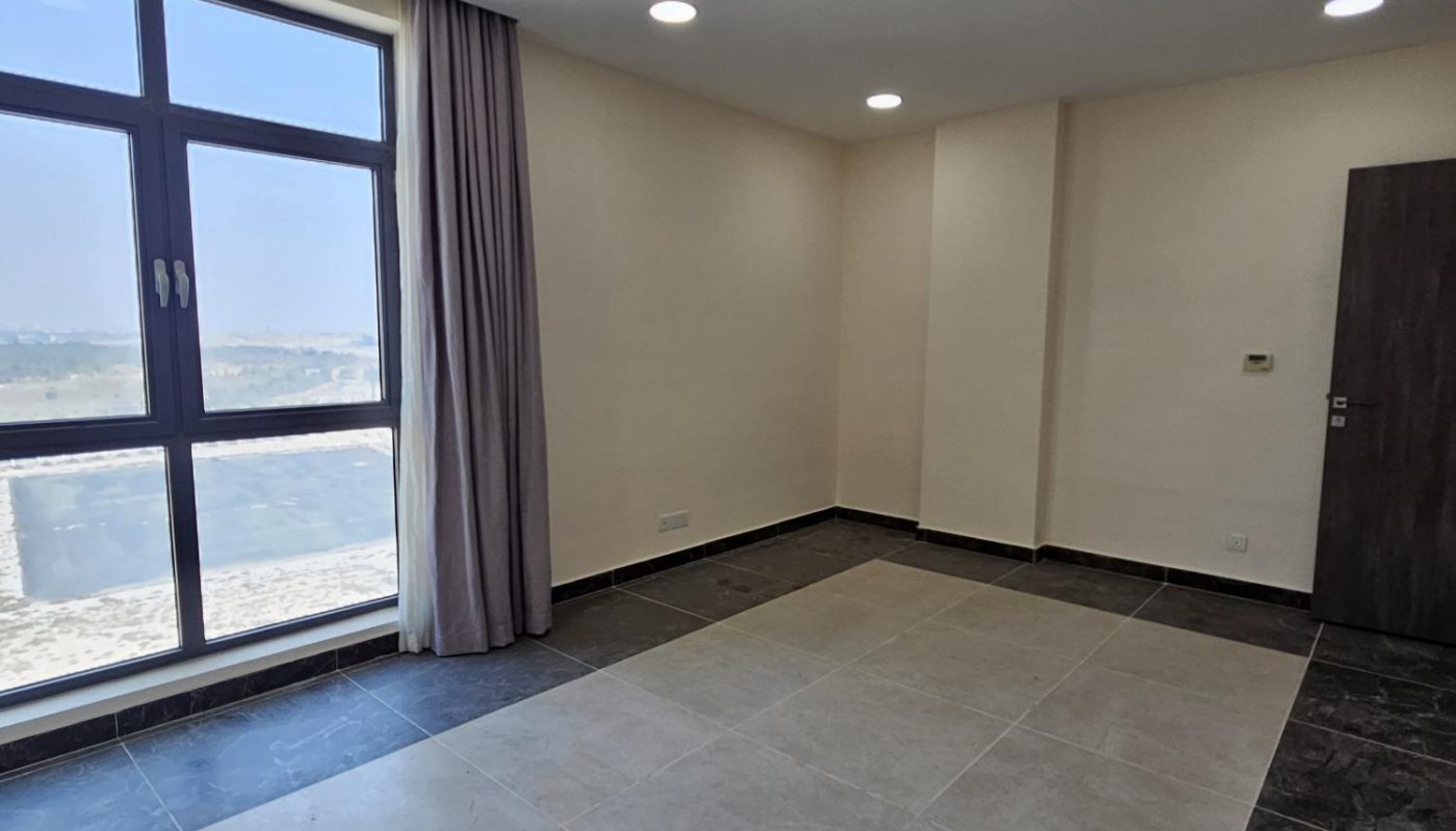 An empty, modern room with tiled floors, cream-colored walls, recessed ceiling lights, a large window with a curtain, and a dark wooden door.