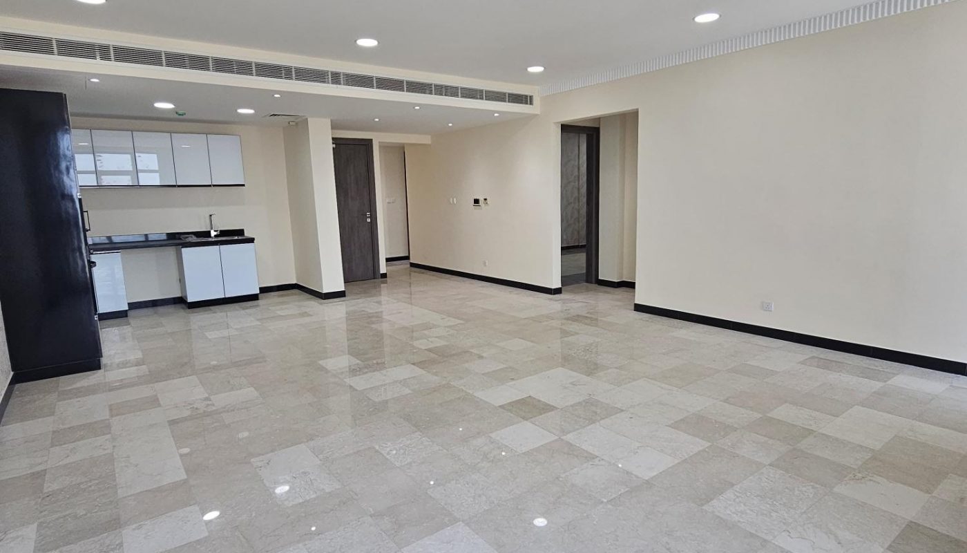 Spacious empty room with tiled floor, white walls, recessed lighting, and an open kitchen area at the far end.