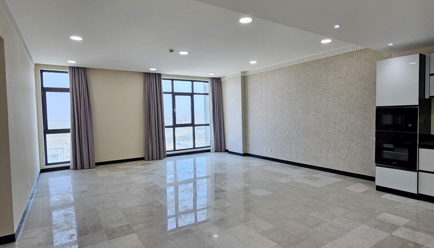 Spacious, empty living room with tiled floors, large windows with beige curtains, white ceiling lights, and a modern built-in kitchen area on the right.