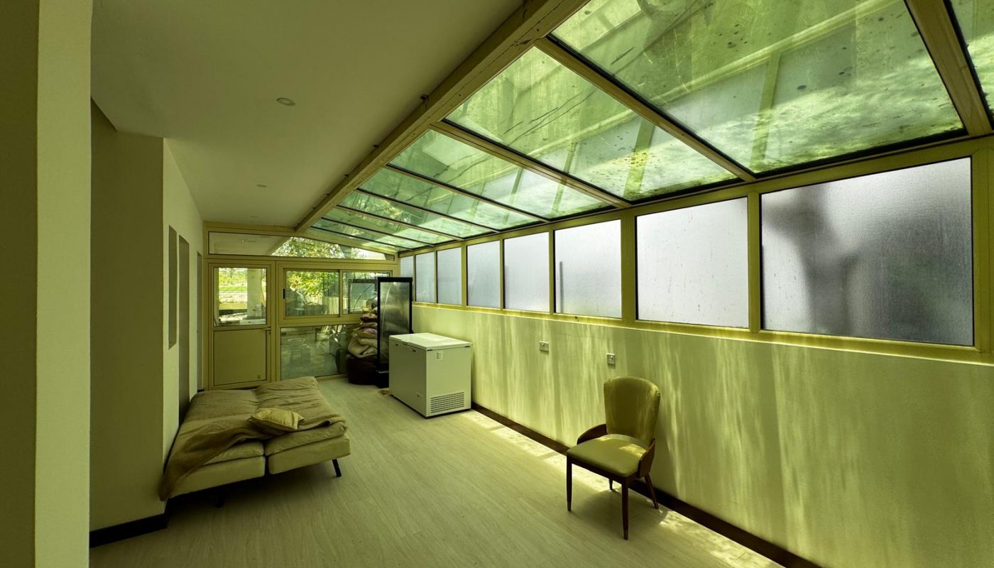 A narrow room with large slanted windows, a single chair, a white chest freezer, and a bed along the left wall; sunlight filters through the glass ceiling.