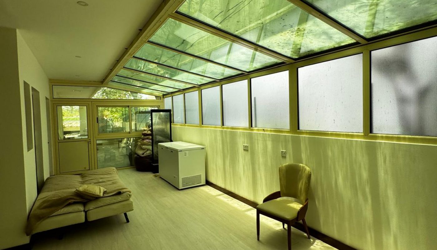 A narrow room with a glass ceiling, a single chair, a sofa, a fridge, and bags stacked in the back. Sunlight streams in through the green-tinted glass roof.
