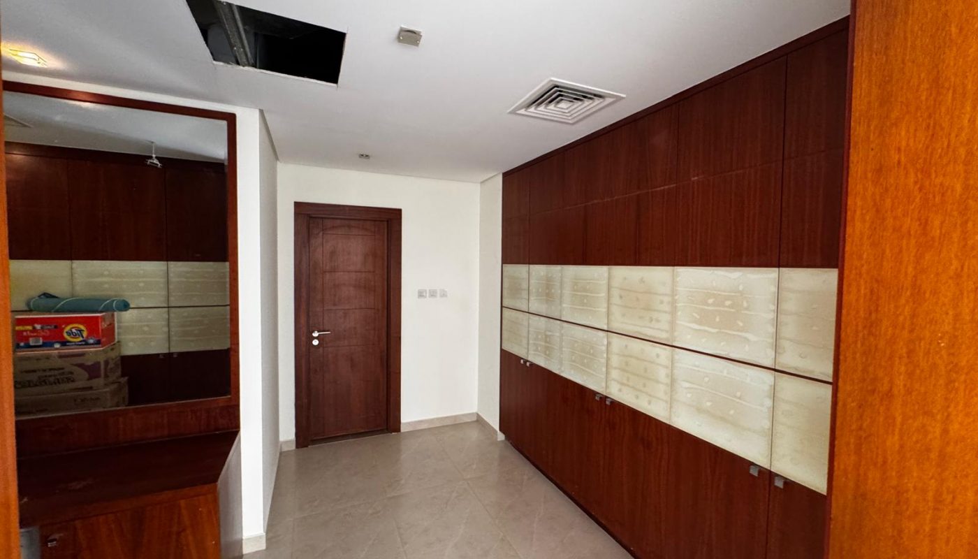 Small room with tiled floor, wooden cabinets along one wall, a mirror, stacked boxes, a closed wooden door, and an open ceiling panel.
