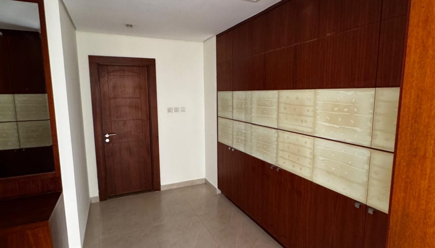 A hallway with beige floor tiles, wooden cabinets along one wall, a closed wooden door, and a ceiling vent. One ceiling panel is missing, exposing an opening.