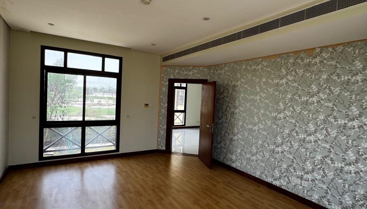 Empty room with wooden flooring, floral-patterned wallpaper on one wall, a large window, and an open door leading to another room. Natural light enters through the window.