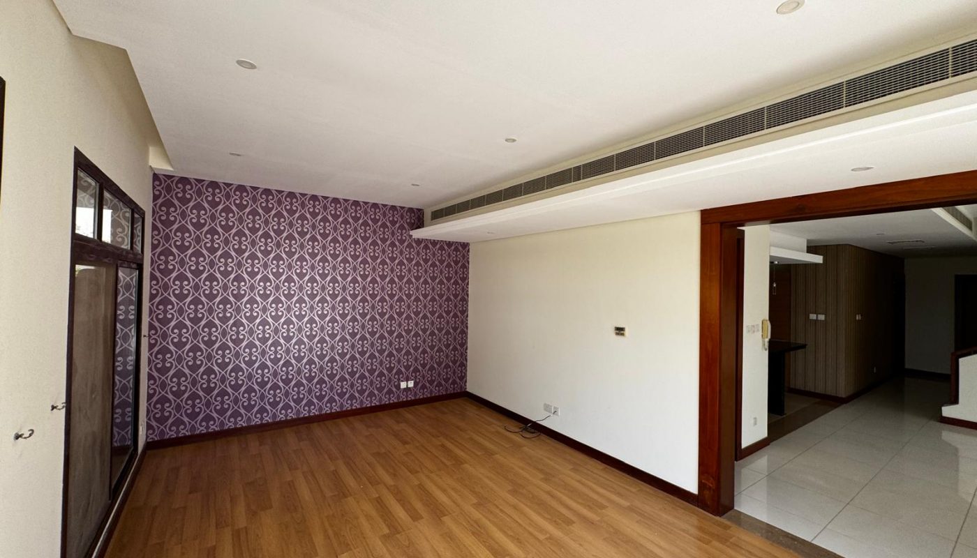 A spacious, unfurnished room with wood flooring, a purple patterned accent wall, white ceiling, and recessed lighting, connected to a tiled hallway with wooden trims.
