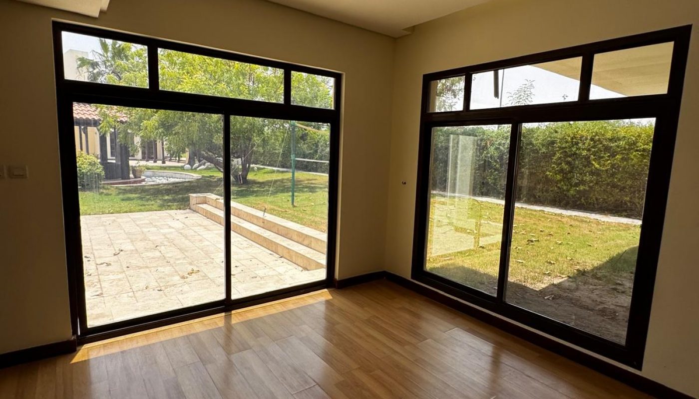 Sunlit room with wooden flooring, large sliding glass doors, and windows overlooking a garden and patio area outside.