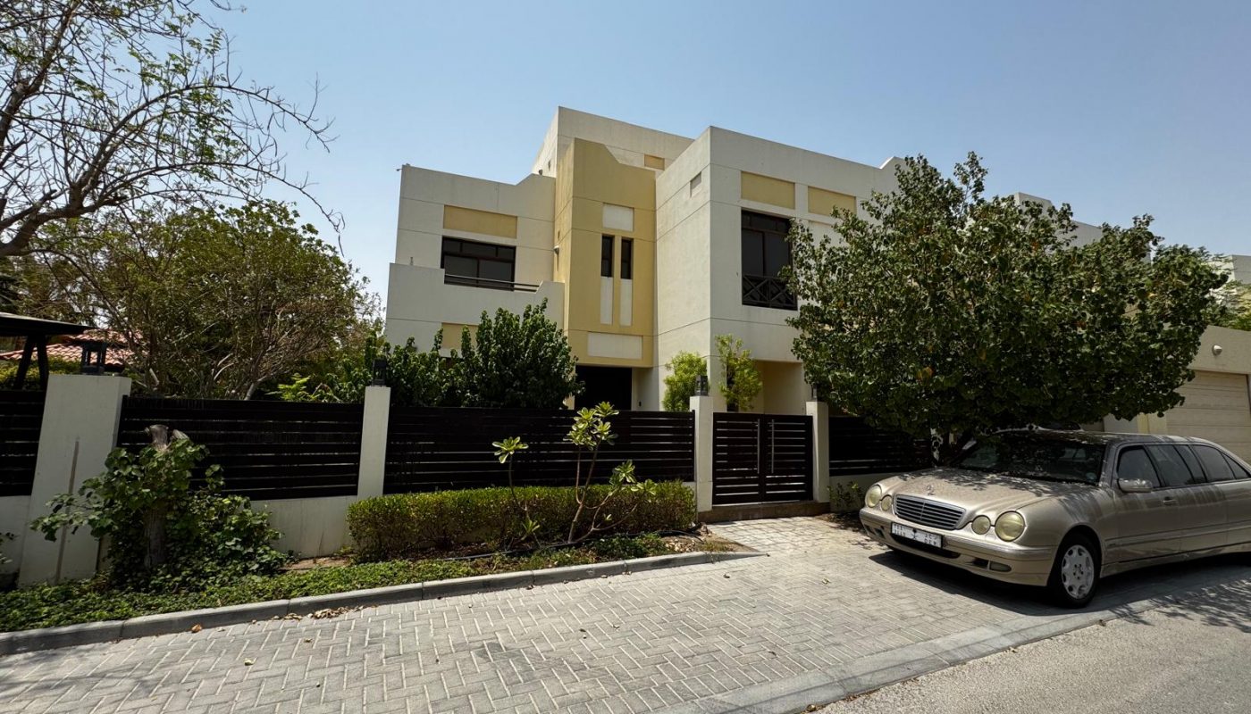 Two-story modern house with beige exterior, large windows, and a dark gated fence. A silver sedan is parked on the street in front of the house. Trees and shrubs line the property.