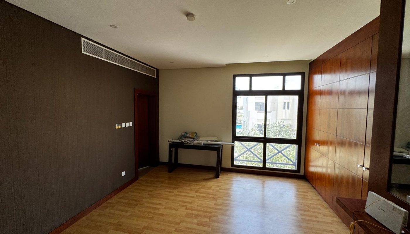 A mostly empty room with wood flooring, a large window, a small table with items on it, dark and light walls, and built-in wood cabinets along one side.