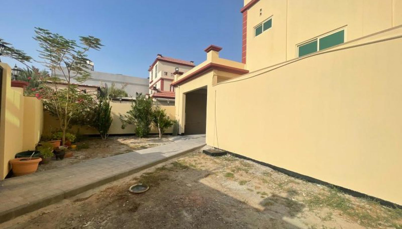 A paved walkway leads to a garage beside a yellow two-story house with green-trimmed windows and a small garden area with potted plants.