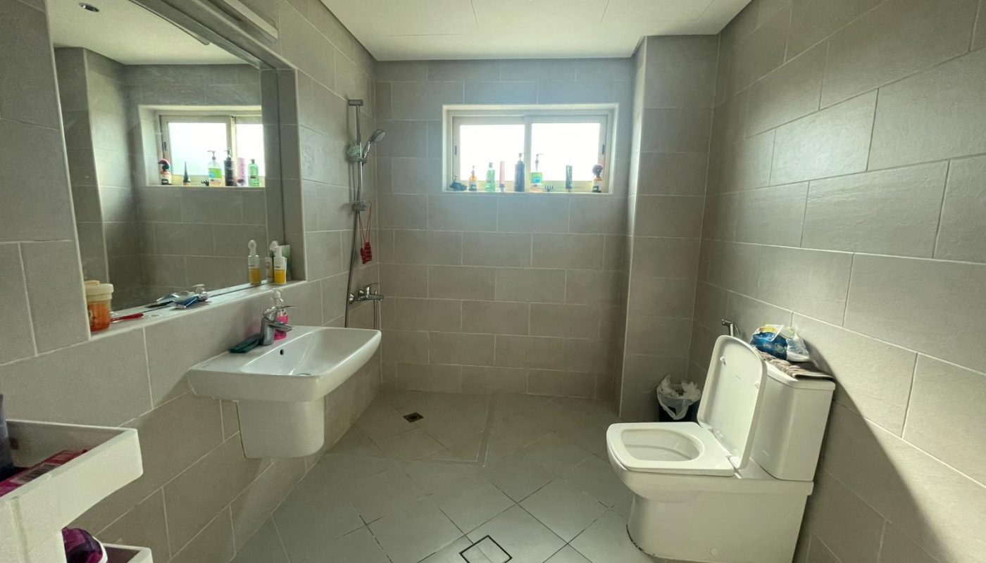 Spacious bathroom with beige tiled walls, a toilet, wall-mounted sink, mirror, shower area, and a window with toiletries on the sill.