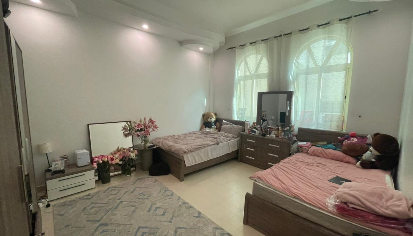 A tidy bedroom with two single beds, plush toys, a rug, flowers in vases, a dresser with a mirror, and large windows with sheer curtains letting in daylight.