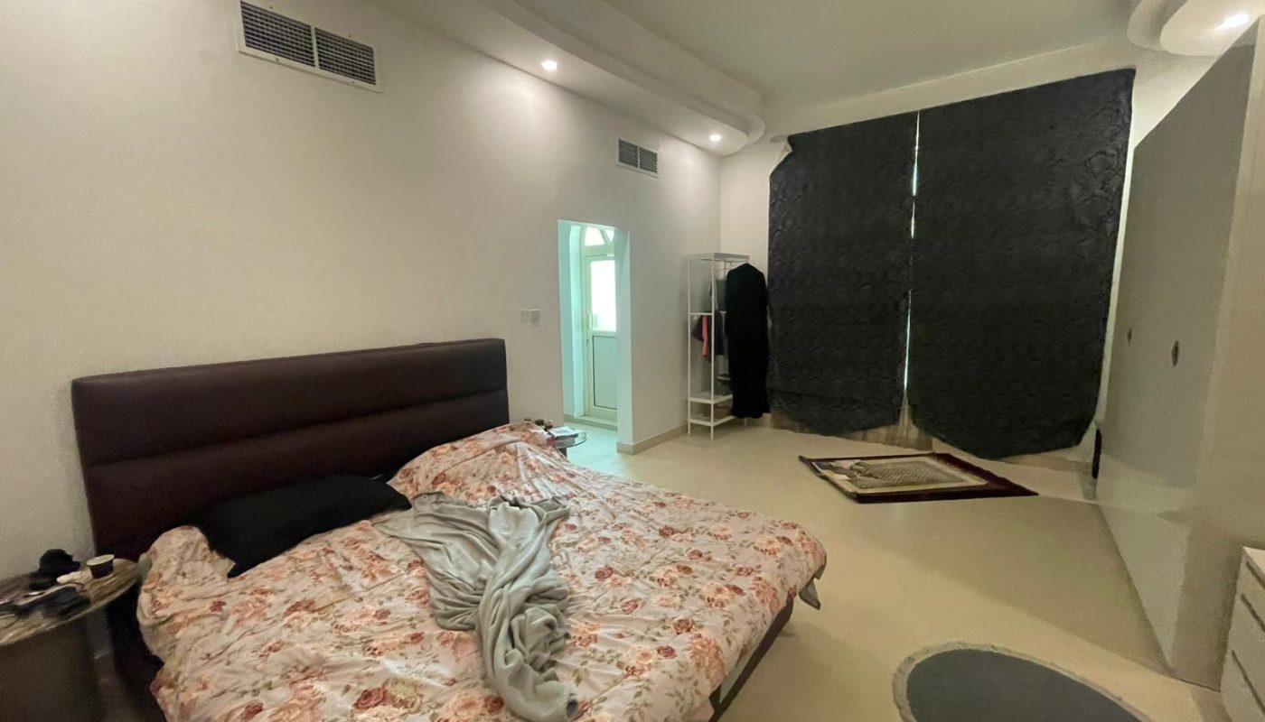 A bedroom with an unmade bed, floral bedding, black pillow, small side table, open wardrobe, dark curtains, and a round rug on a light-colored floor.