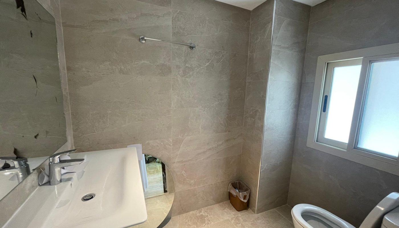 A bathroom with a sink, mirror, towel rack, toilet, small waste bin, and frosted window; the walls and floor are tiled in beige.