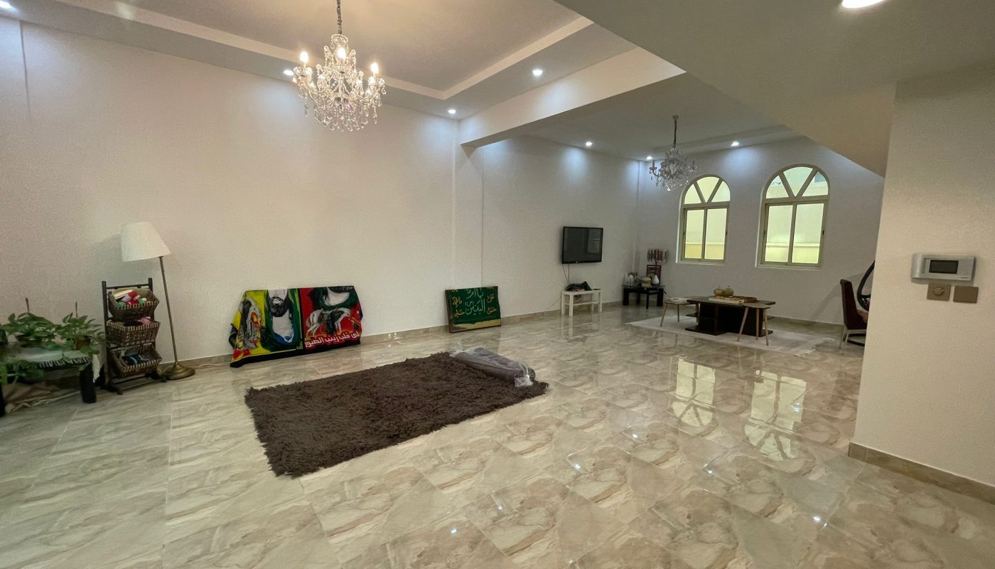 Spacious living room with marble floors, a brown rug, chandeliers, wall art on the floor, plants, and a seating area with a TV and arched windows in the background.