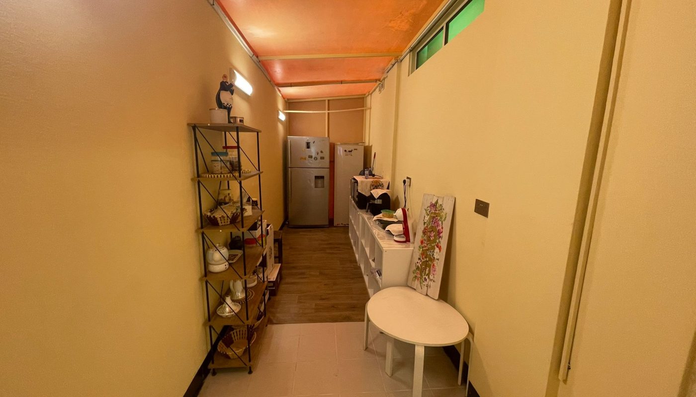 Narrow indoor hallway with shelving on the left, kitchen counters on the right, a round table, and a refrigerator at the far end. Warm lighting illuminates the space.
