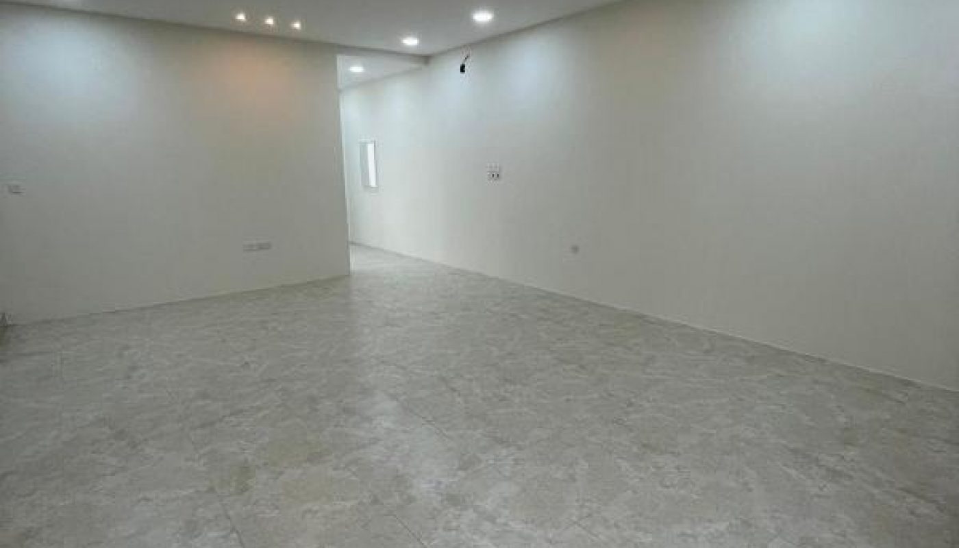 Spacious empty room with light-colored tiled floor, white walls, and multiple recessed ceiling lights. No furniture or decorations present.