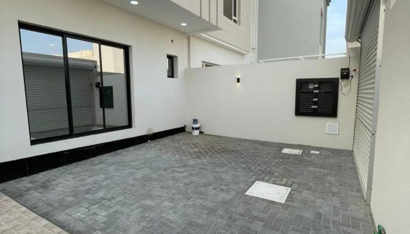 A modern house exterior with a paved courtyard, large window, sliding gate, utility meters, and tiled flooring under a partly cloudy sky.