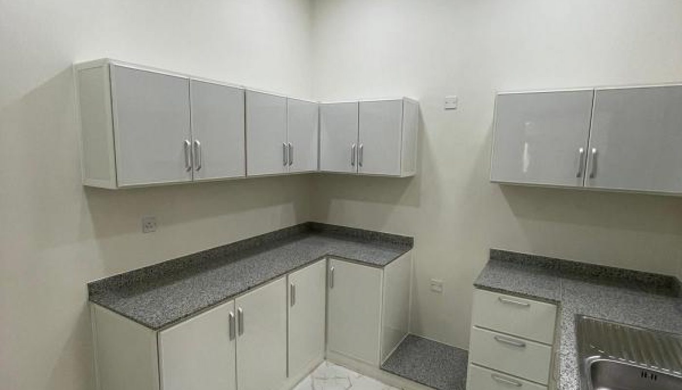 A modern kitchen with white cabinets, granite countertops, and a stainless steel sink, featuring white marble-patterned floor tiles and recessed ceiling lights.