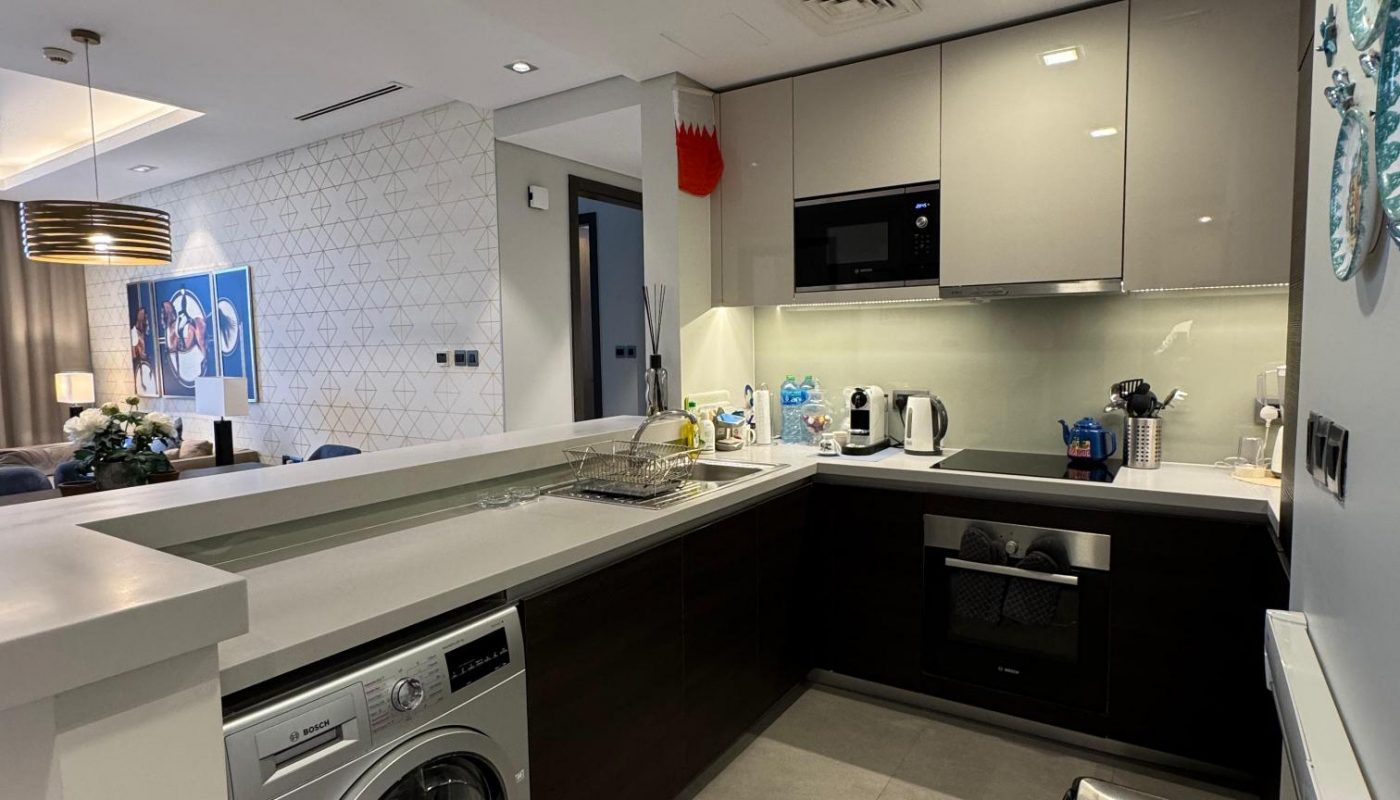Modern kitchen with dark lower cabinets, glossy upper cabinets, sink, countertop appliances, washing machine, trash bin, and partial view of dining and living area.