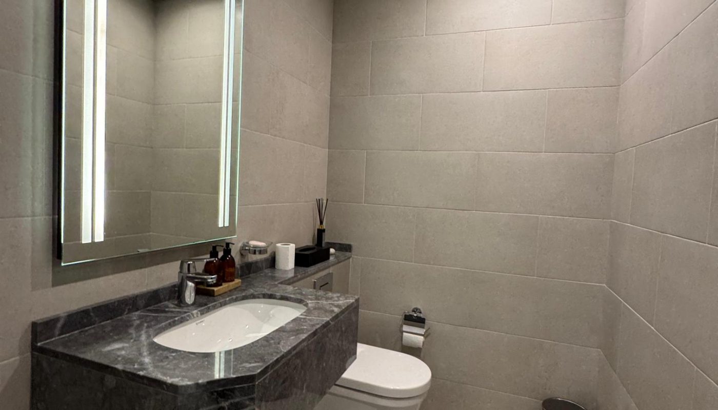 Modern bathroom with light gray tiles, a wall-mounted toilet, a floating sink with a marble countertop, a large mirror, and minimal accessories. A trash bin and rugs are on the floor.