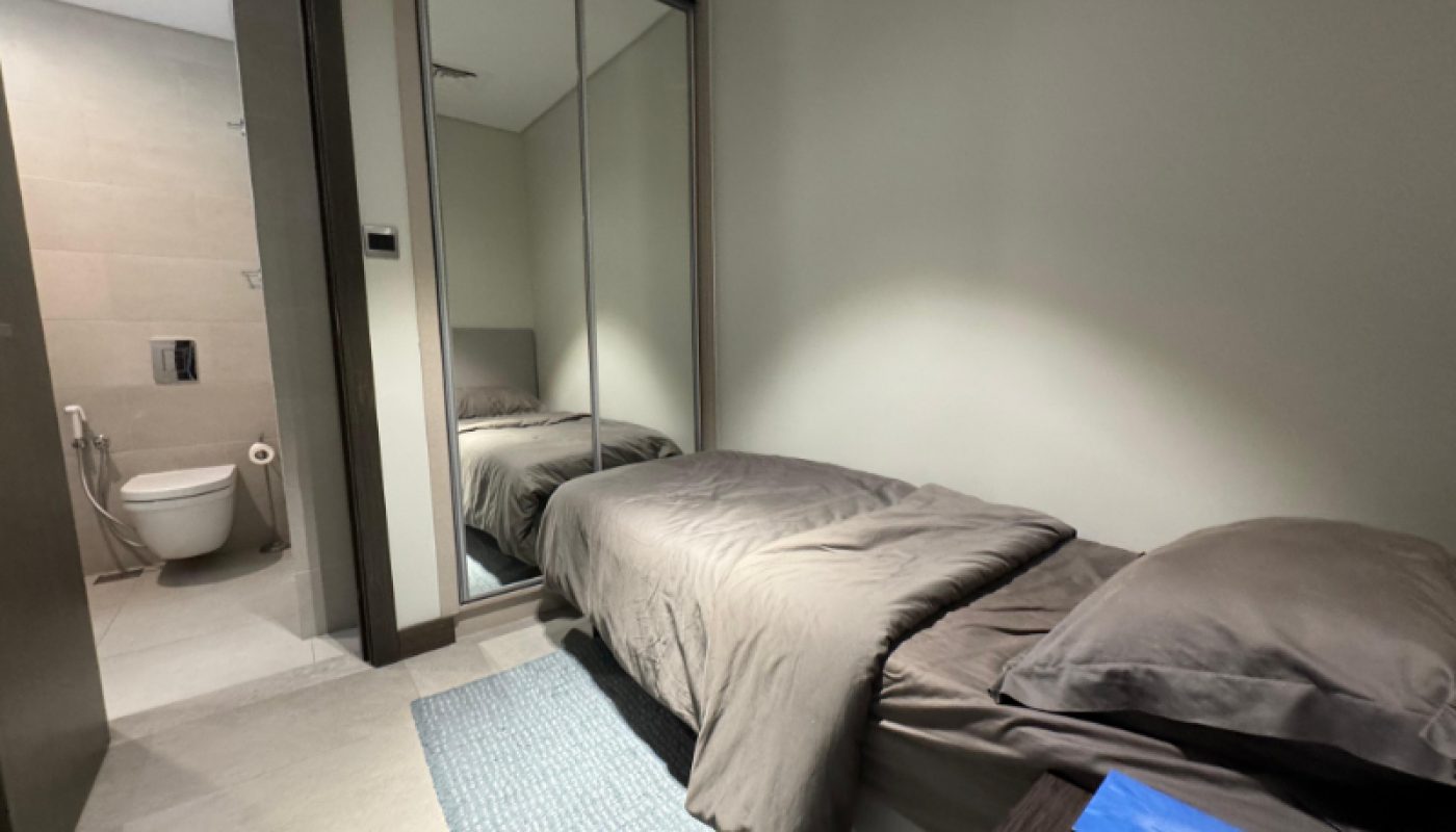A small, neatly made bed with gray bedding is next to a mirrored closet, with an adjacent bathroom visible through an open door.