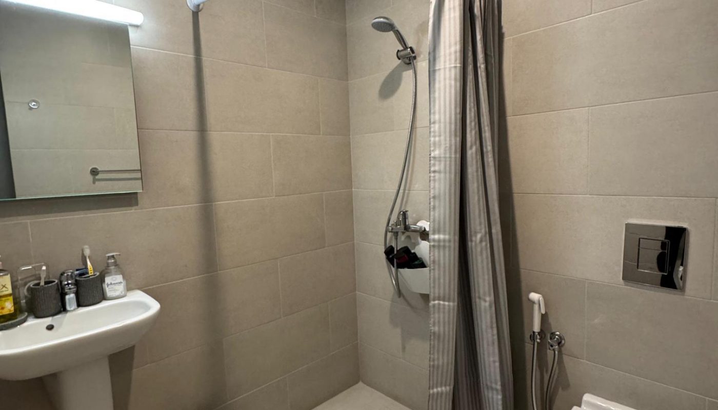 A modern bathroom with a wall-mounted sink, mirror, trash bin, shower area with a curtain, handheld showerhead, and a wall-mounted toilet with a bidet spray.