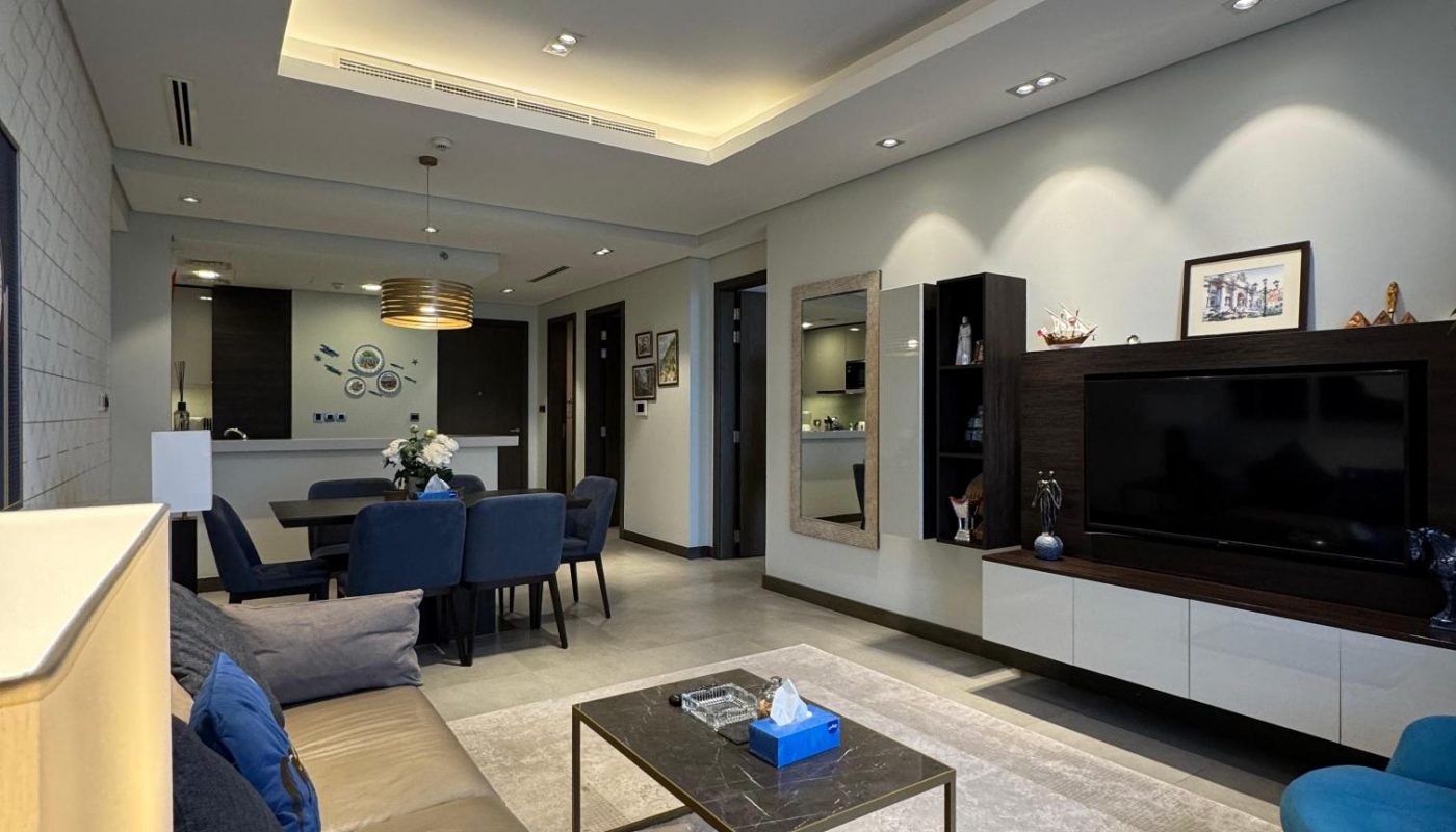 Modern living room with neutral tones, blue accents, a wall-mounted TV, built-in shelving, and a dining area visible in the background.