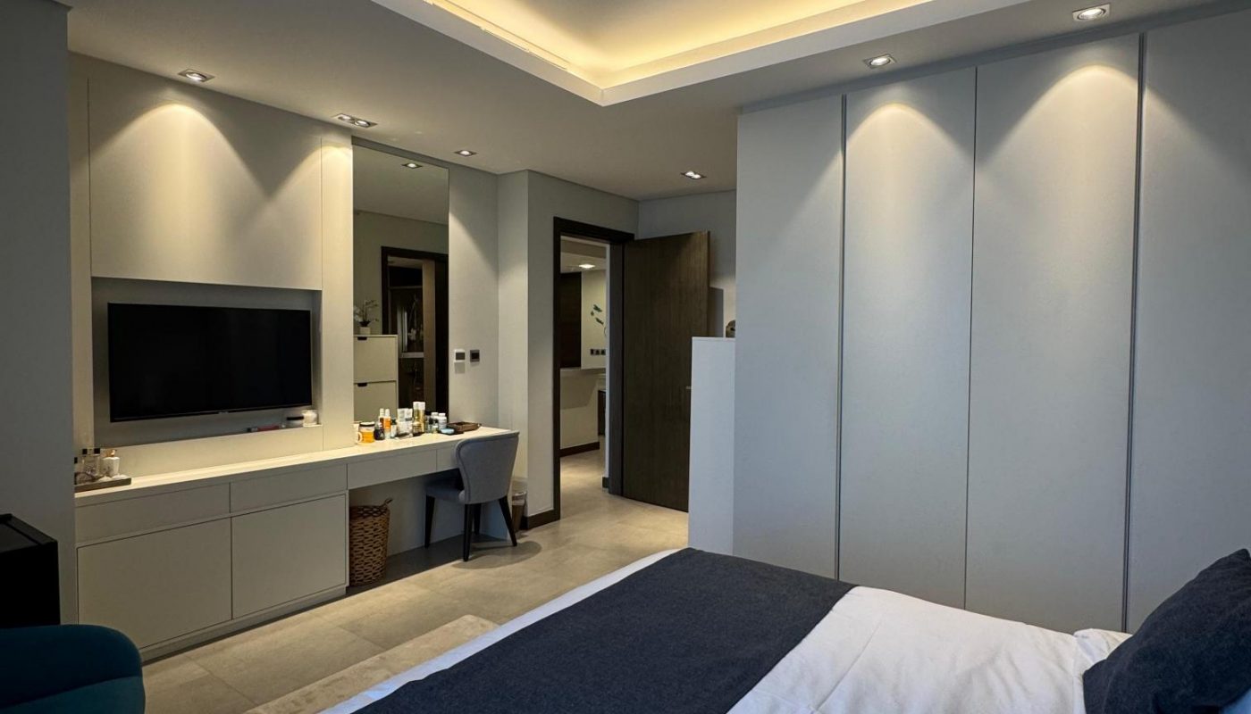 Modern bedroom with a double bed, dark blue bedding, wall-mounted TV, desk area, and recessed ceiling lighting. Room features neutral tones and minimalistic decor.