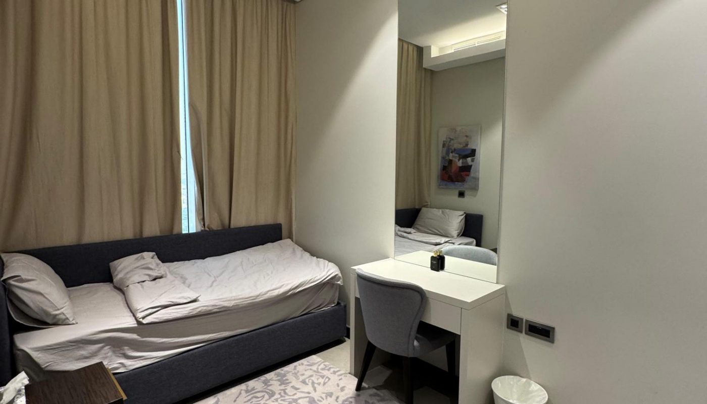 A modern bedroom with a single bed, a large mirror above a white desk and chair, beige curtains, a patterned rug, and a small waste bin.