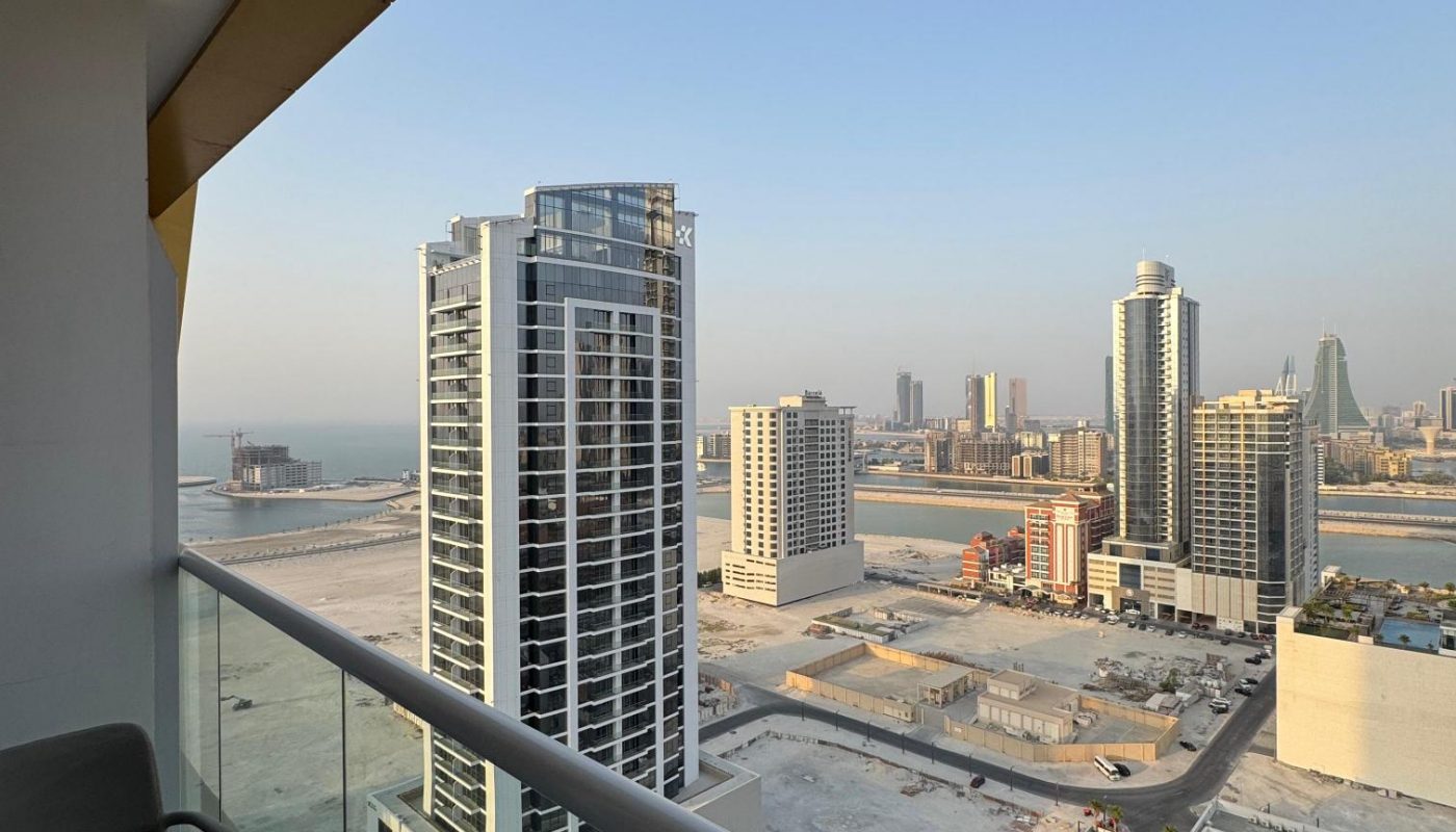 View from a high-rise balcony overlooking modern buildings, empty lots, and a distant sea under a clear sky in an urban area.