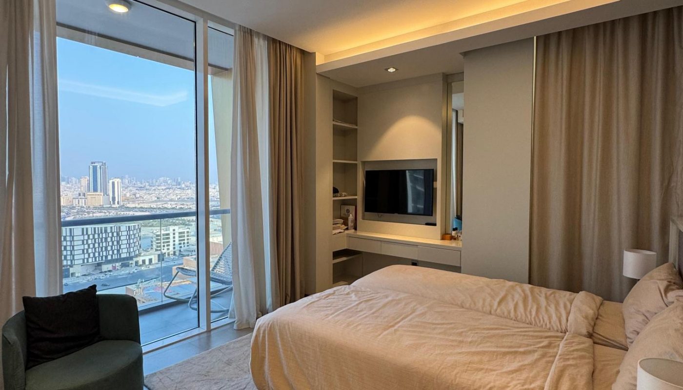 Modern bedroom with beige bedding, a wall-mounted TV, armchair, and large glass doors leading to a balcony with a city view.
