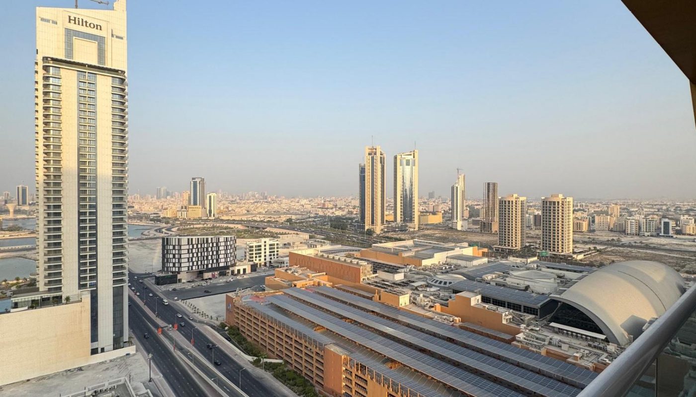 Cityscape view from a high-rise balcony showing modern buildings, a Hilton hotel, roads, and clear sky during daylight.