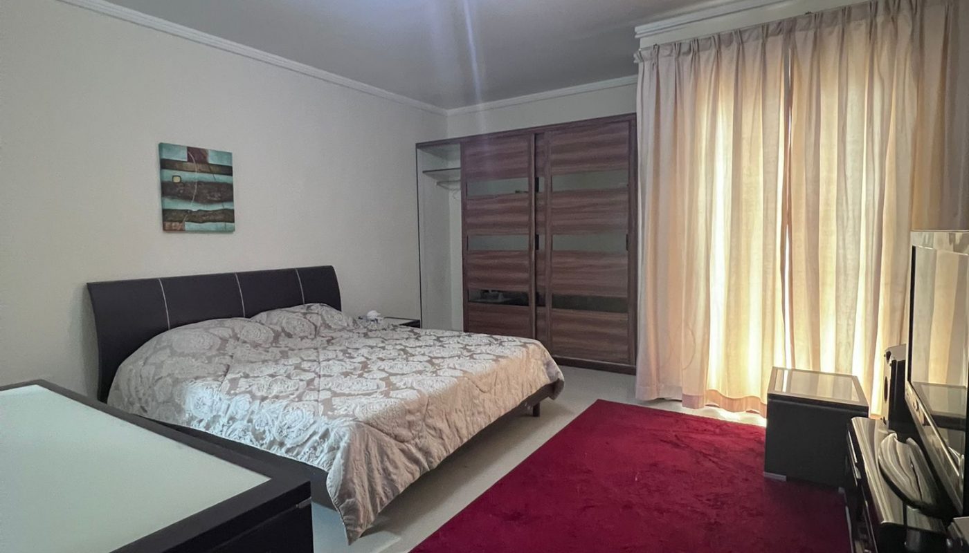 A bedroom with a double bed, a red rug, a wardrobe with sliding doors, a set of drawers, and a window covered by beige curtains.
