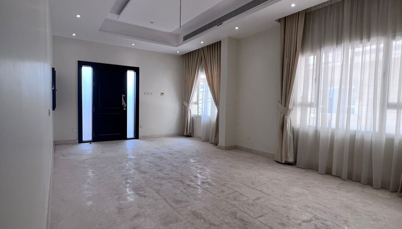 Spacious empty room with marble floors, recessed ceiling lights, large windows with sheer curtains, and a dark front door.