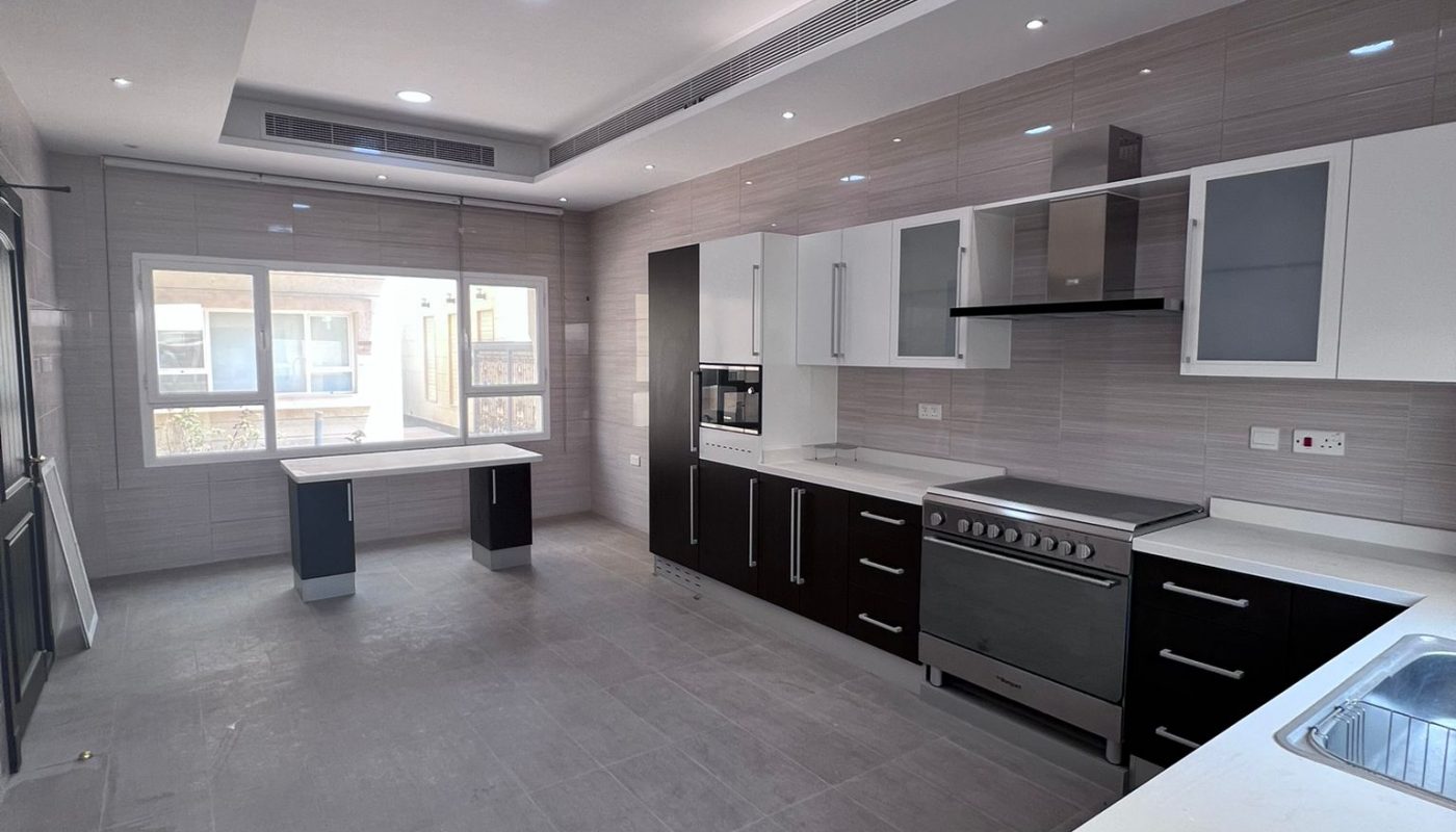 Modern kitchen with gray tile floor, built-in appliances, dark cabinets, white countertops, and under-cabinet lighting. Large windows and recessed ceiling lights provide ample illumination.