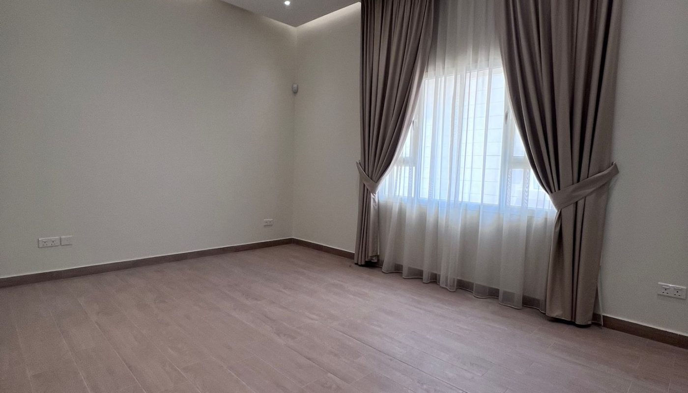 Empty room with light-colored wooden flooring, plain off-white walls, ceiling spotlights, and a window with sheer white curtains and brown drapes.