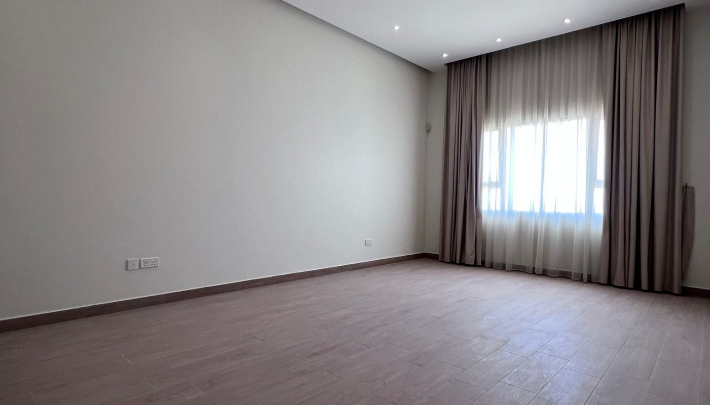 Spacious empty room with light-colored walls, brown tile flooring, ceiling spotlights, and large window covered by sheer curtains and dark drapes.