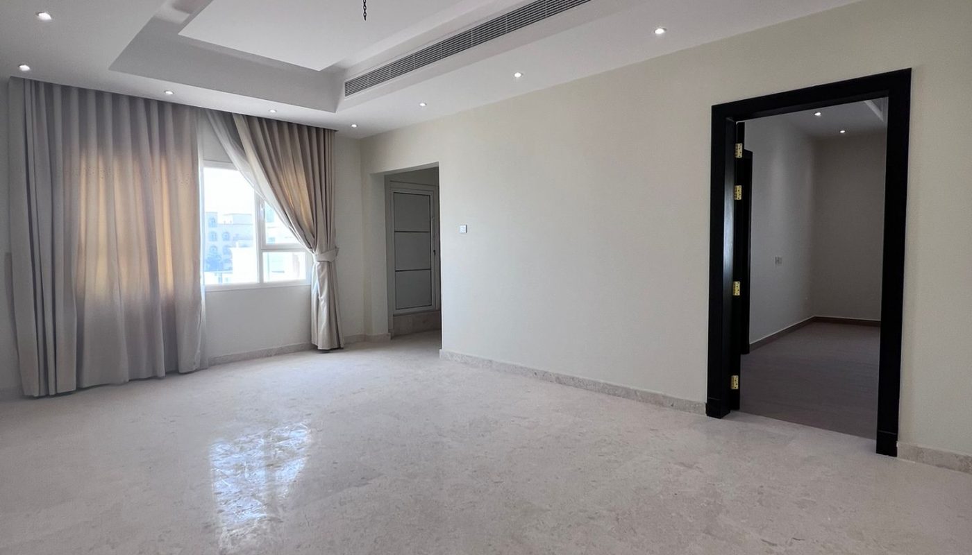 Spacious empty room with marble flooring, white walls, recessed ceiling lights, a large window with curtains, and an open doorway leading to another area.