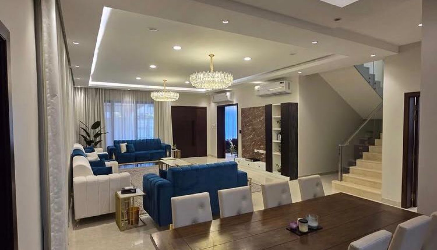 Modern open-plan living and dining room with blue sofas, a wooden dining table with white chairs, chandeliers, and a staircase leading upstairs.