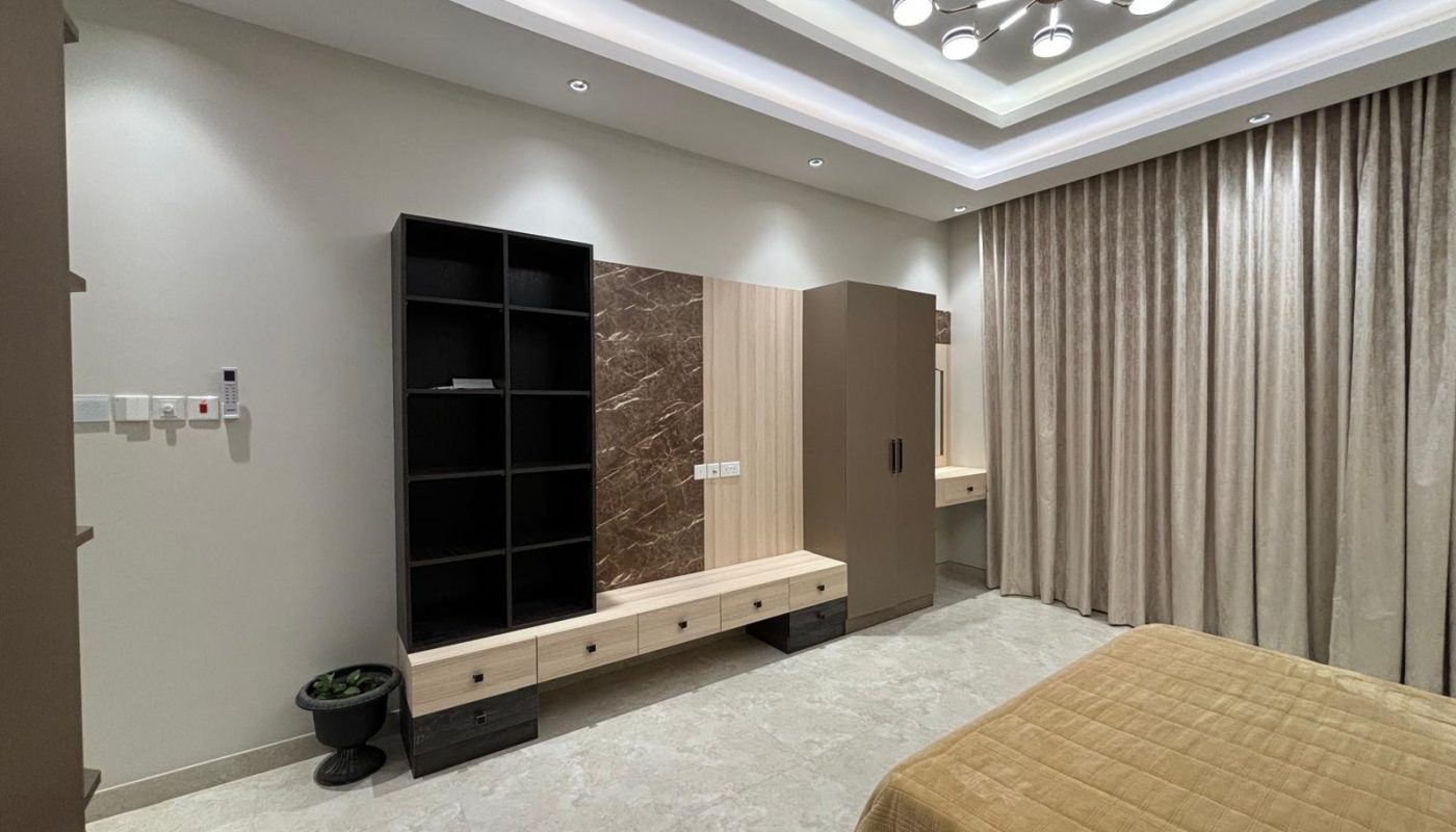 Modern bedroom with beige walls, marble flooring, built-in wooden shelves, a large mirror, ceiling lights, and floor-length curtains. Part of a bed is visible in the foreground.