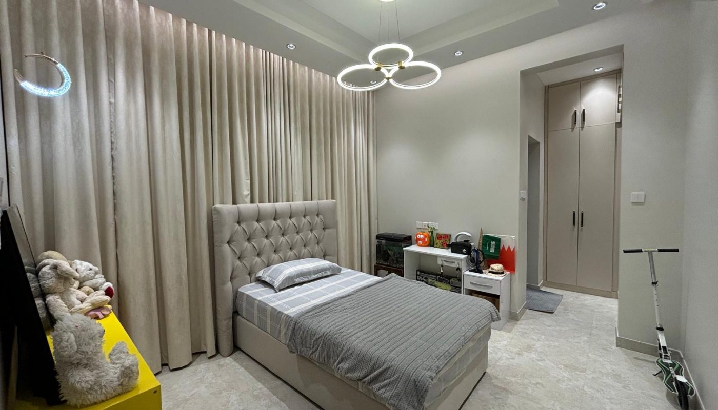 A modern bedroom with a single bed, grey bedding, neutral walls, a yellow nightstand with stuffed animals, and a contemporary ceiling light fixture.