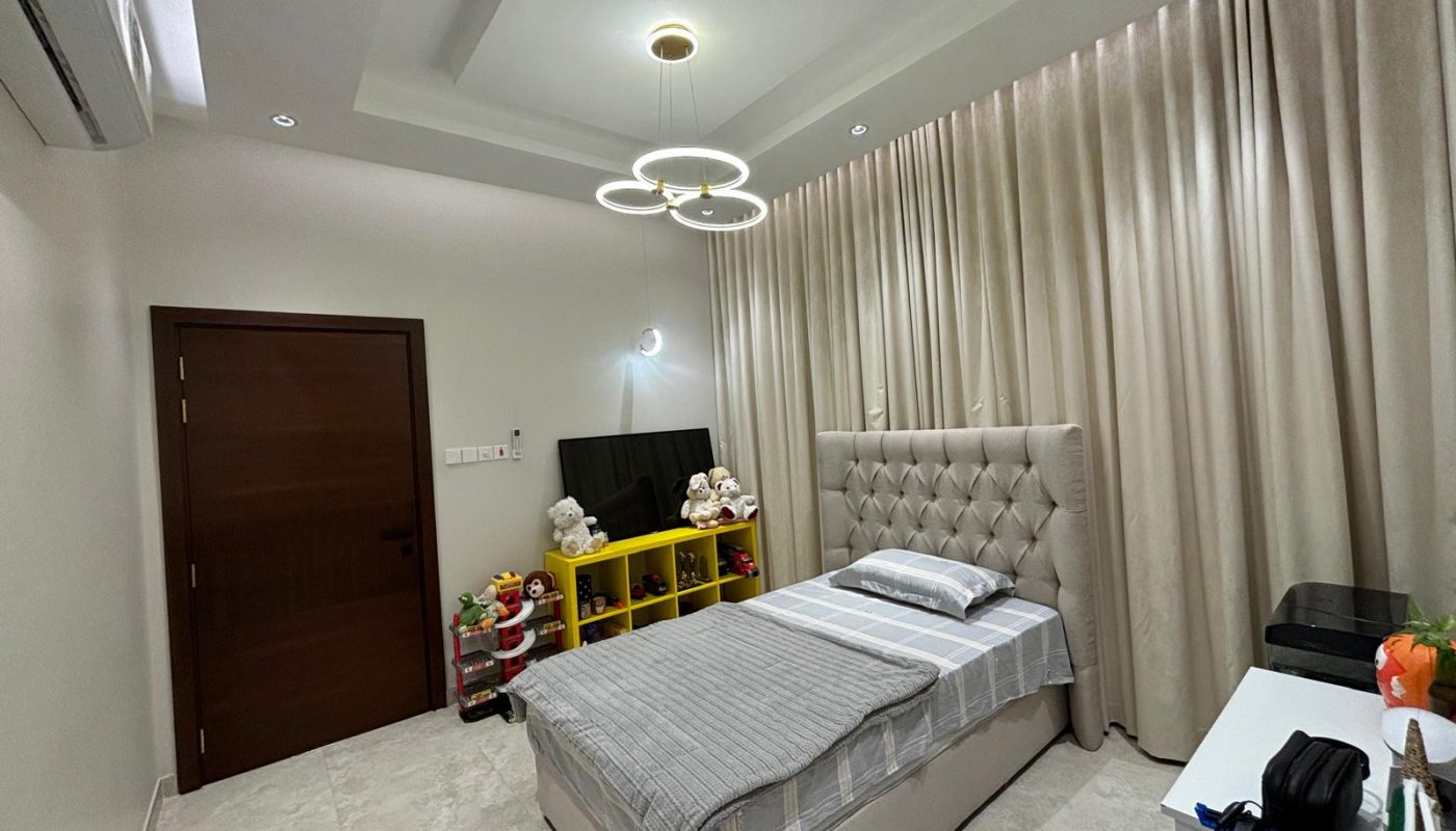 A neatly made single bed in a modern bedroom with cream walls, a tufted headboard, a TV on a yellow shelf, toys, and beige curtains covering the window.