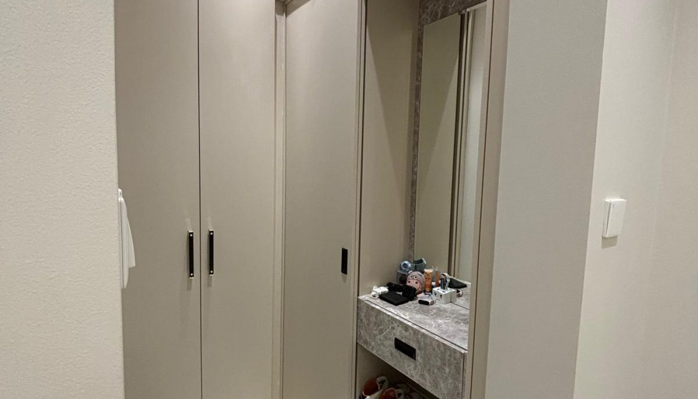 A modern hallway with beige built-in cabinets, a mirror, open shelves holding shoes, and a small countertop with various personal items.