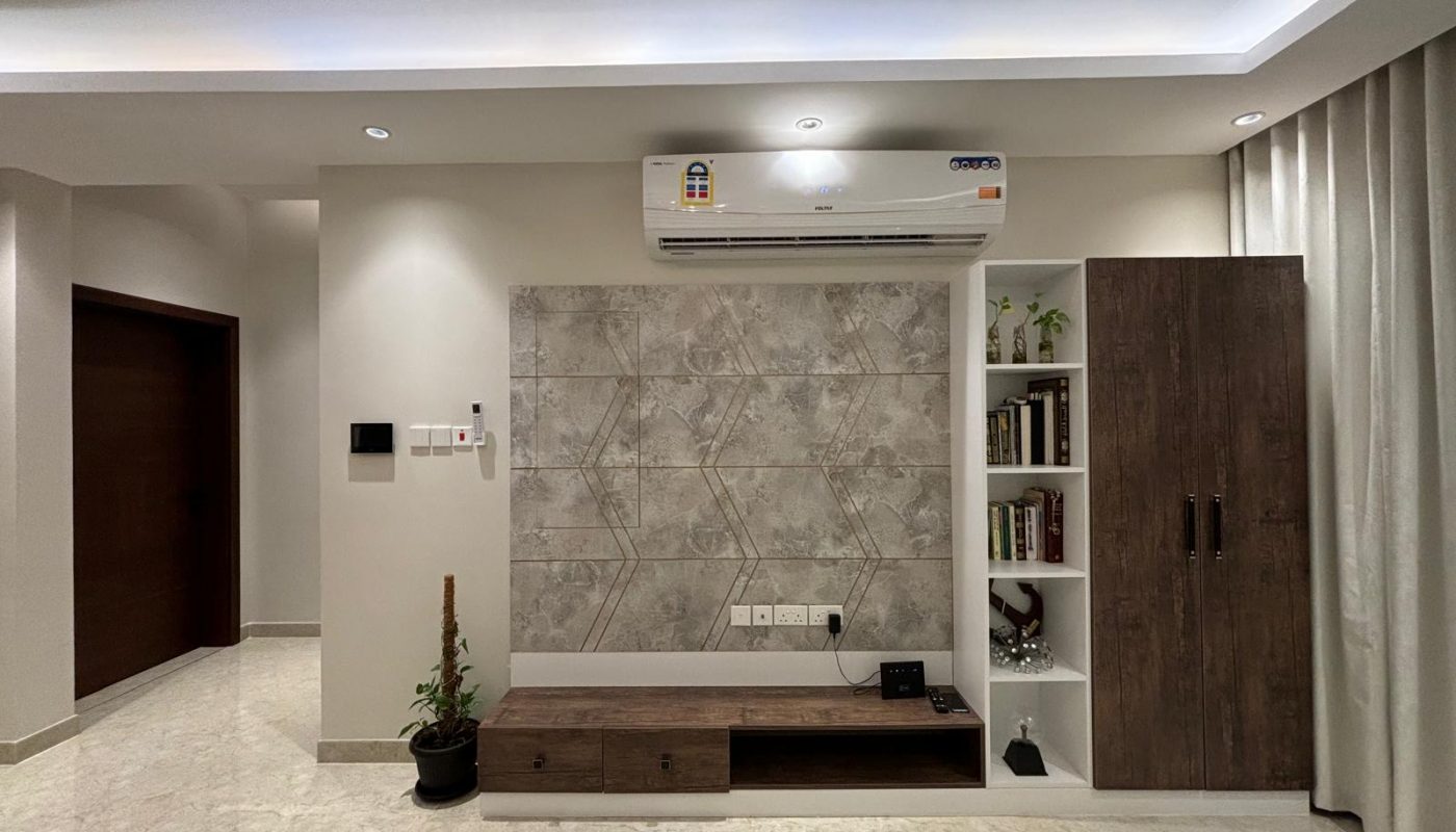 Modern living room with a wall-mounted air conditioner, tile accent wall, wooden TV unit, shelving with books and decor, potted plant, and beige flooring.