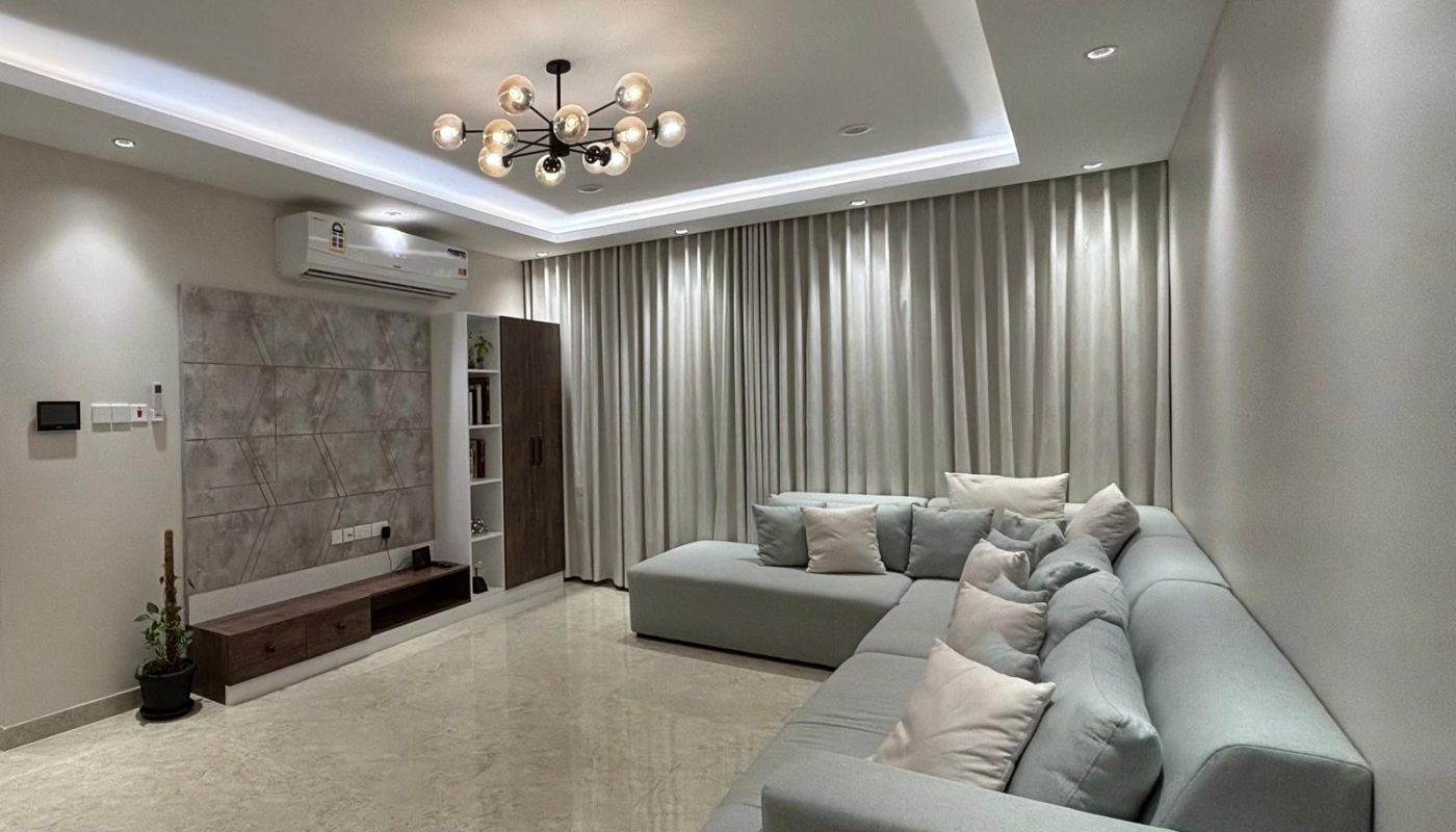 Modern living room with light marble floors, a large L-shaped grey sofa with cushions, a low wooden TV unit, beige curtains, recessed ceiling lights, and a contemporary chandelier.