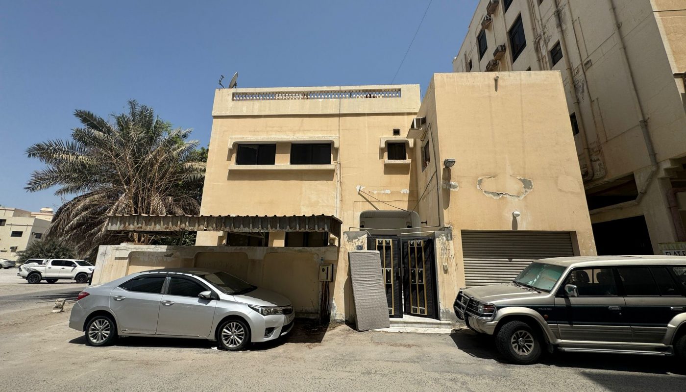 A beige two-story building with a metal gate and awning, two parked cars, a palm tree on the left, and taller buildings nearby under a clear sky.