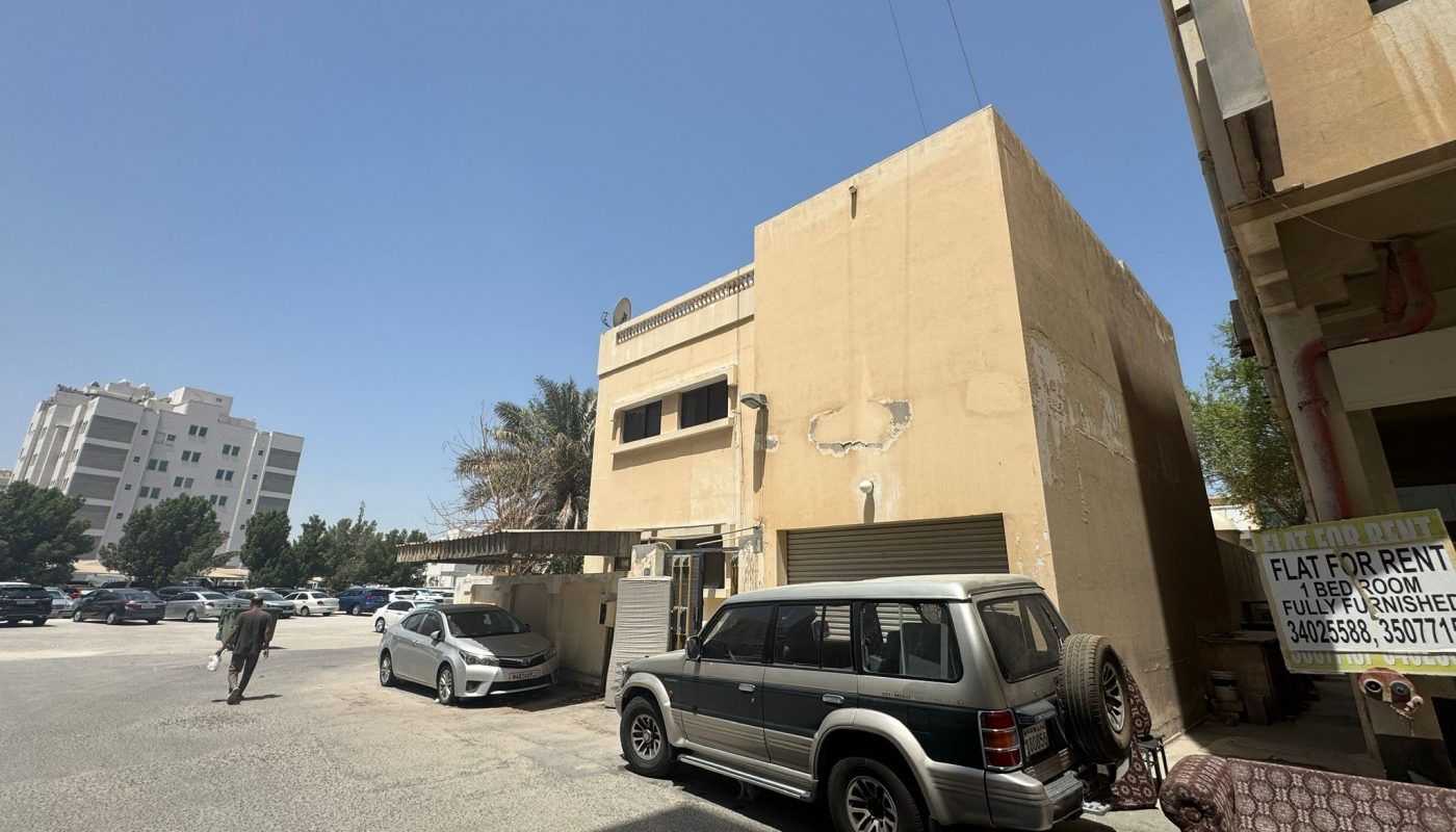 A beige building with a "Flat For Rent" sign, parked cars, and a person walking on a sunny day in an urban area.
