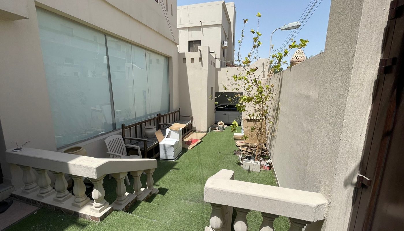 A narrow outdoor area with artificial grass, two benches, a few potted plants, and scattered boxes next to a building with large windows.