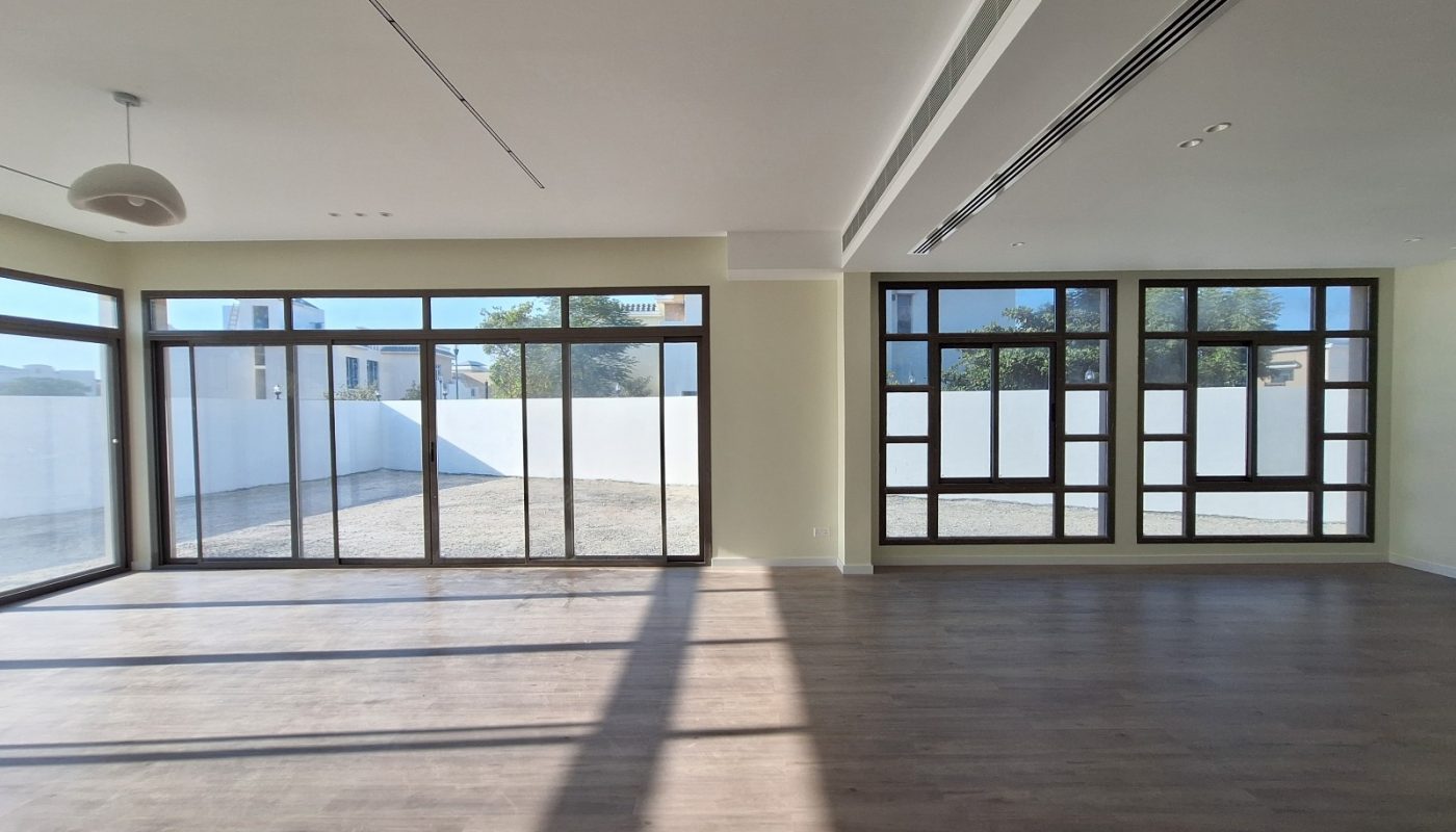 A spacious, empty room with large floor-to-ceiling windows, wood flooring, and sunlight streaming in, overlooking a fenced outdoor area.